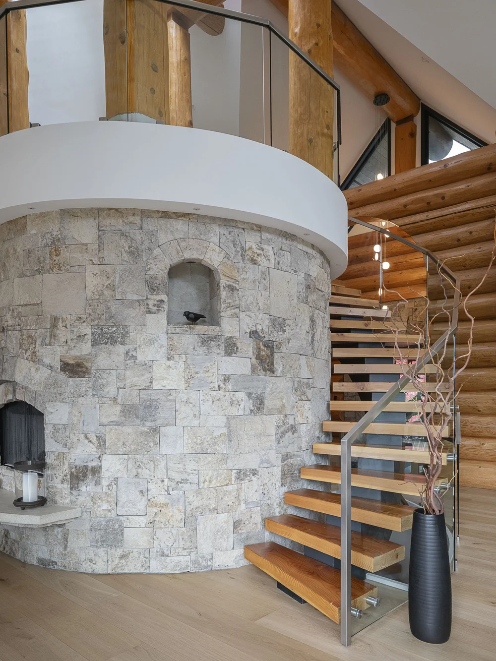 Open tread stair winding around a curved stone wall, glass and steel rail, logs behind