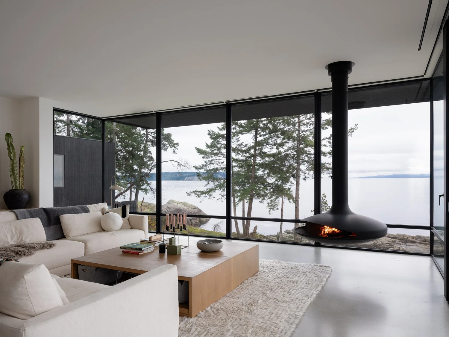 Living room with a pale sectional and low table, the fireplace hanging beside full height glazing
