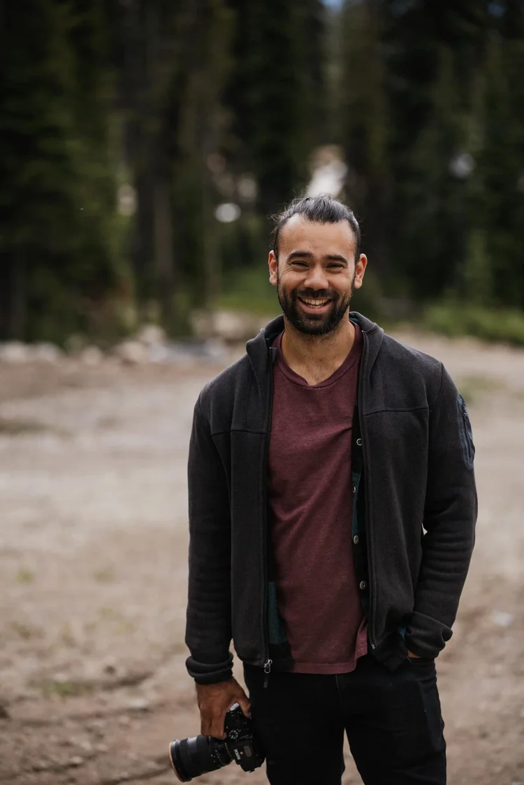 Matt Anthony standing on a gravel clearing holding a camera, conifers behind him