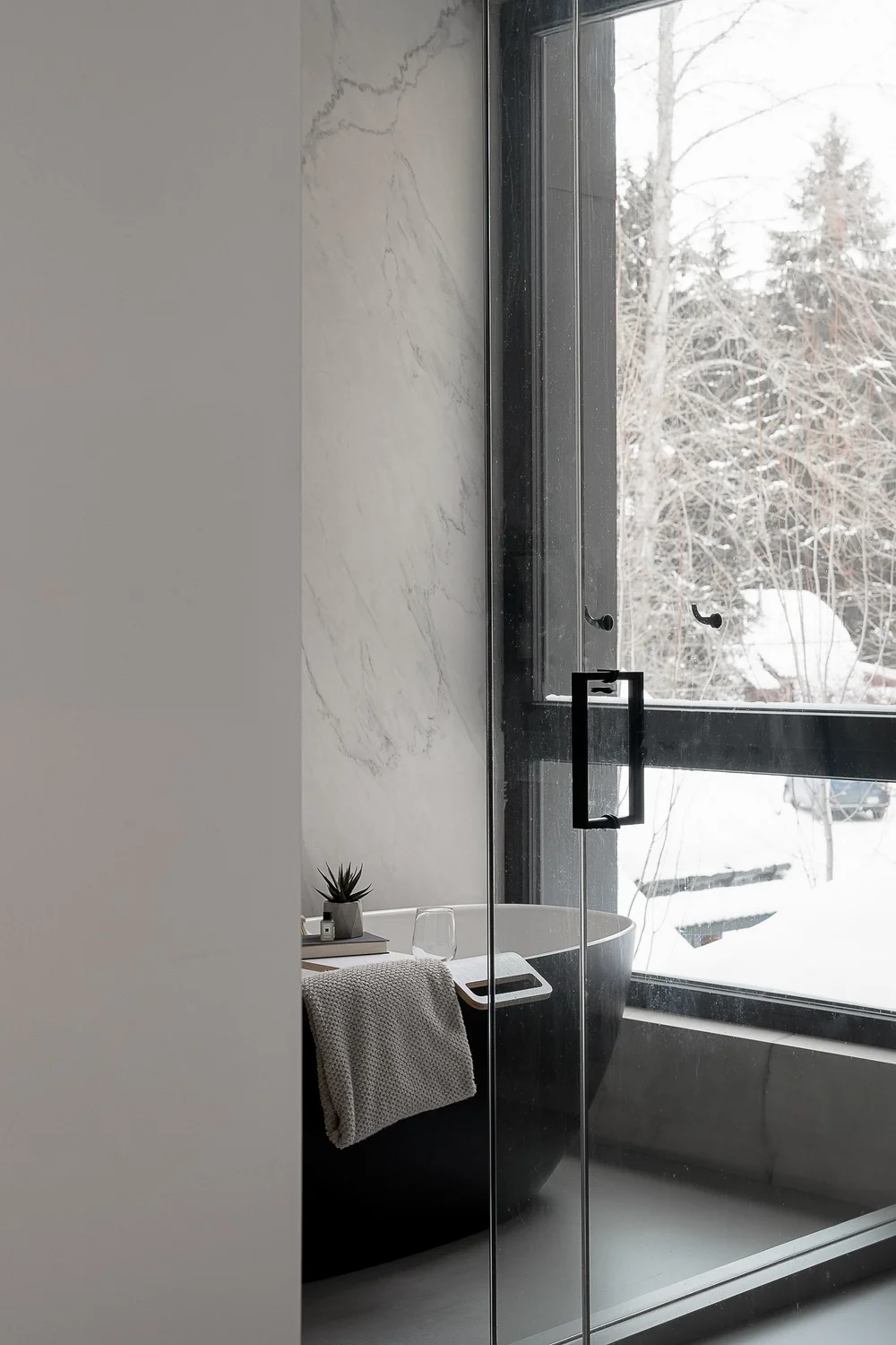 Freestanding tub at a full height window, veined stone wall and a glass shower door