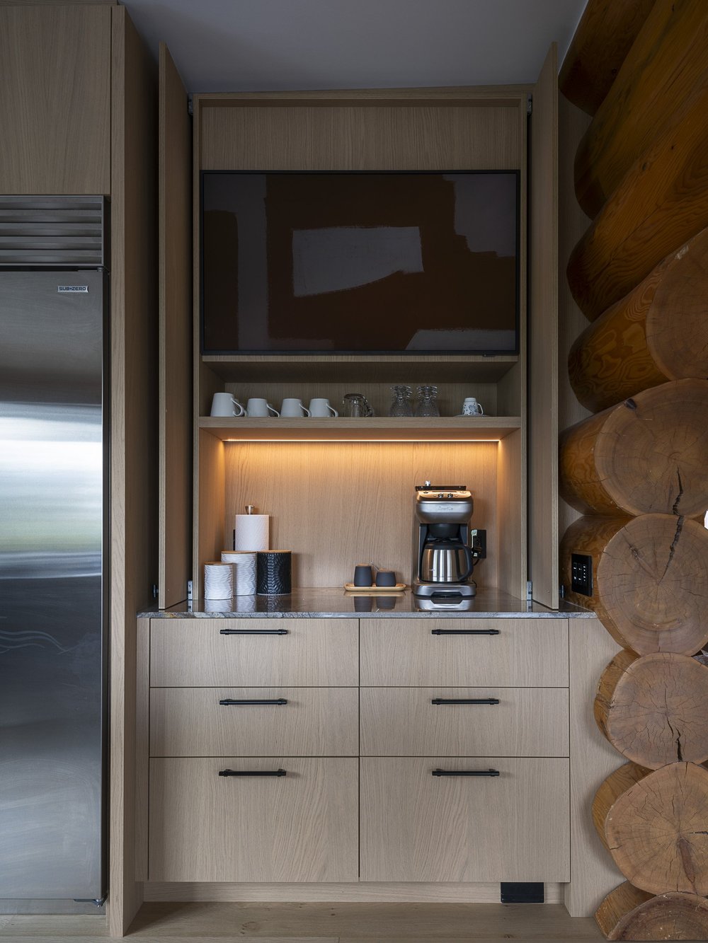 Beverage cabinet standing open, mugs and glasses on a lit shelf below a dark screen