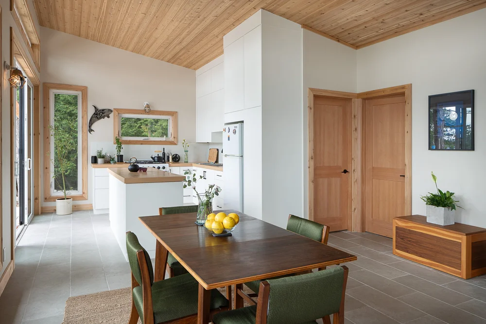 Dining table with green chairs, kitchen island and wood doors under a wood ceiling