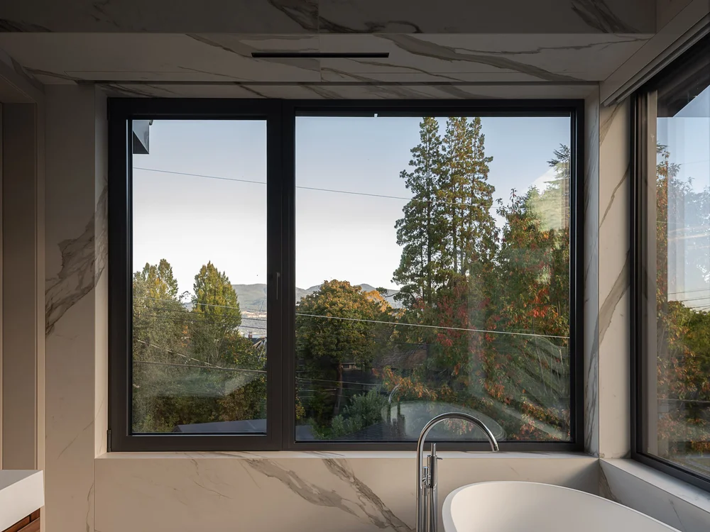 Corner window above the tub in veined stone, trees and a far hillside beyond