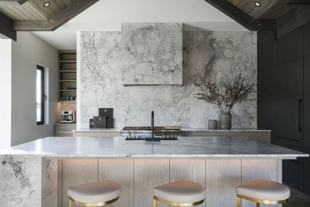 Kitchen island in pale wood with three stools, veined stone wall behind the range