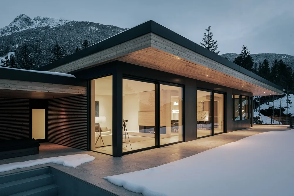 The house at dusk, interiors lit behind full height glazing under a deep timber soffit
