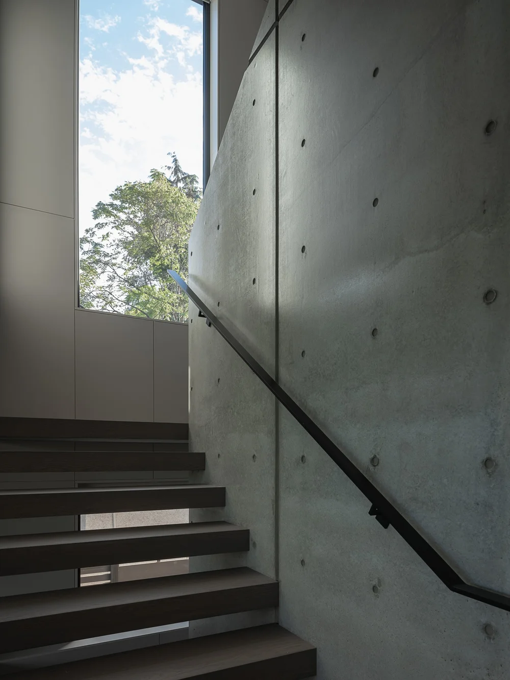 Stair rising beside the concrete wall, a black handrail and a window at the turn