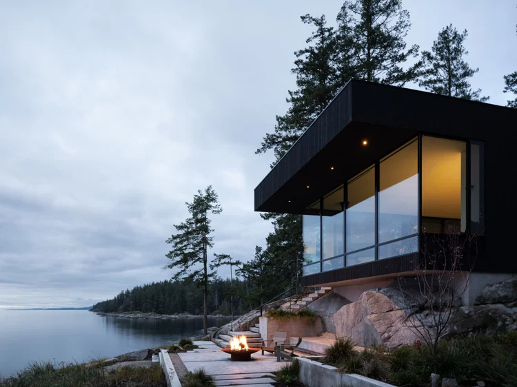 Dark clad house cantilevered over a rock outcrop above the water at dusk, fire bowl on the terrace