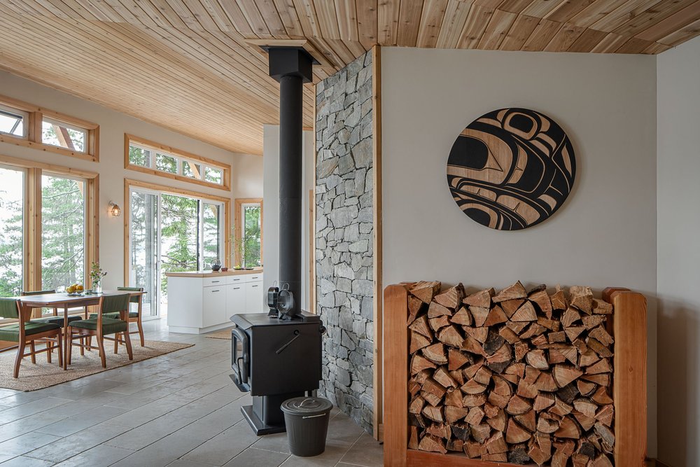 A wood stove against a stone chimney wall, dining table and kitchen beyond