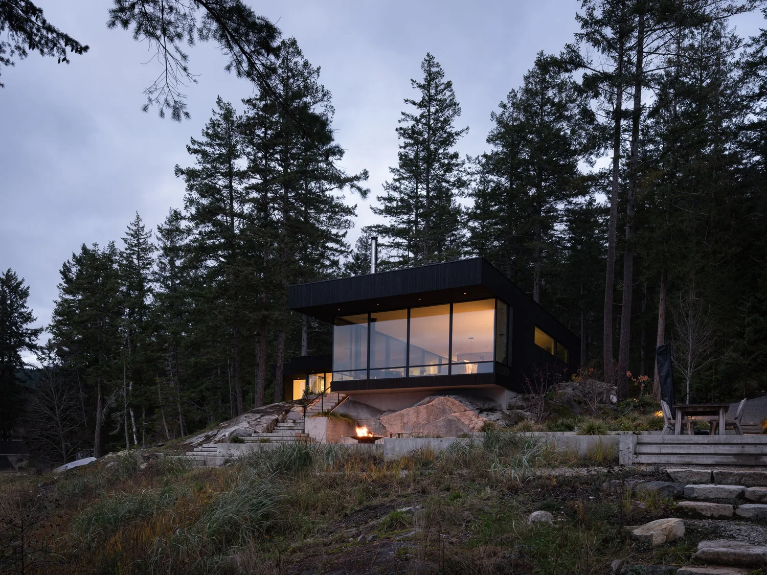 The house at dusk above the rock, warm light behind the glass and a fire lit below