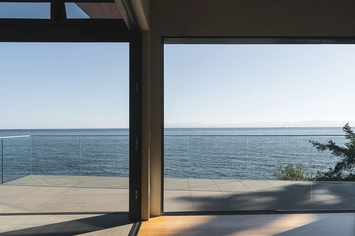 Interior looking out through open sliding glass walls to a paved terrace, a glass guardrail and open water