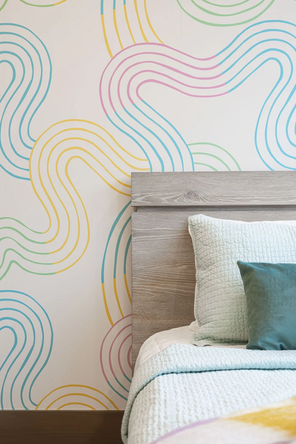 Bed with a timber headboard against a wall of looping coloured lines, a yellow throw