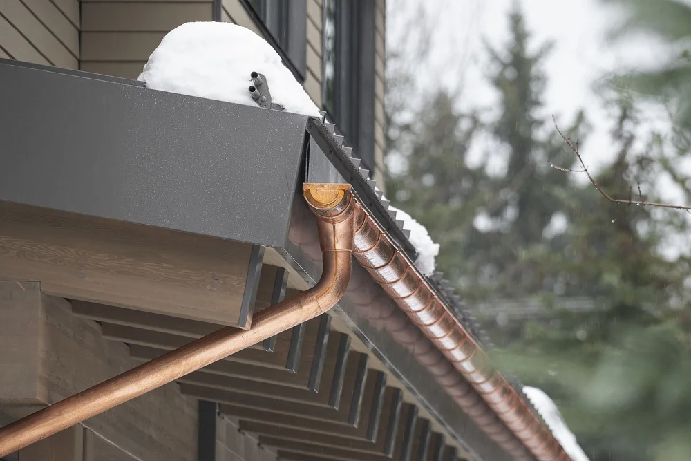 Copper gutter running along a snow covered roof edge, conifers behind