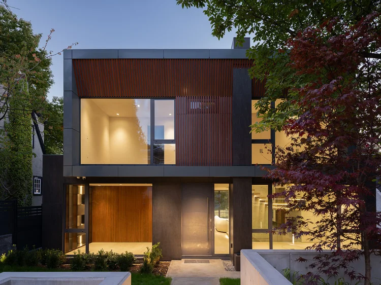 The house at dusk, warm light through the glazing and a red maple at right