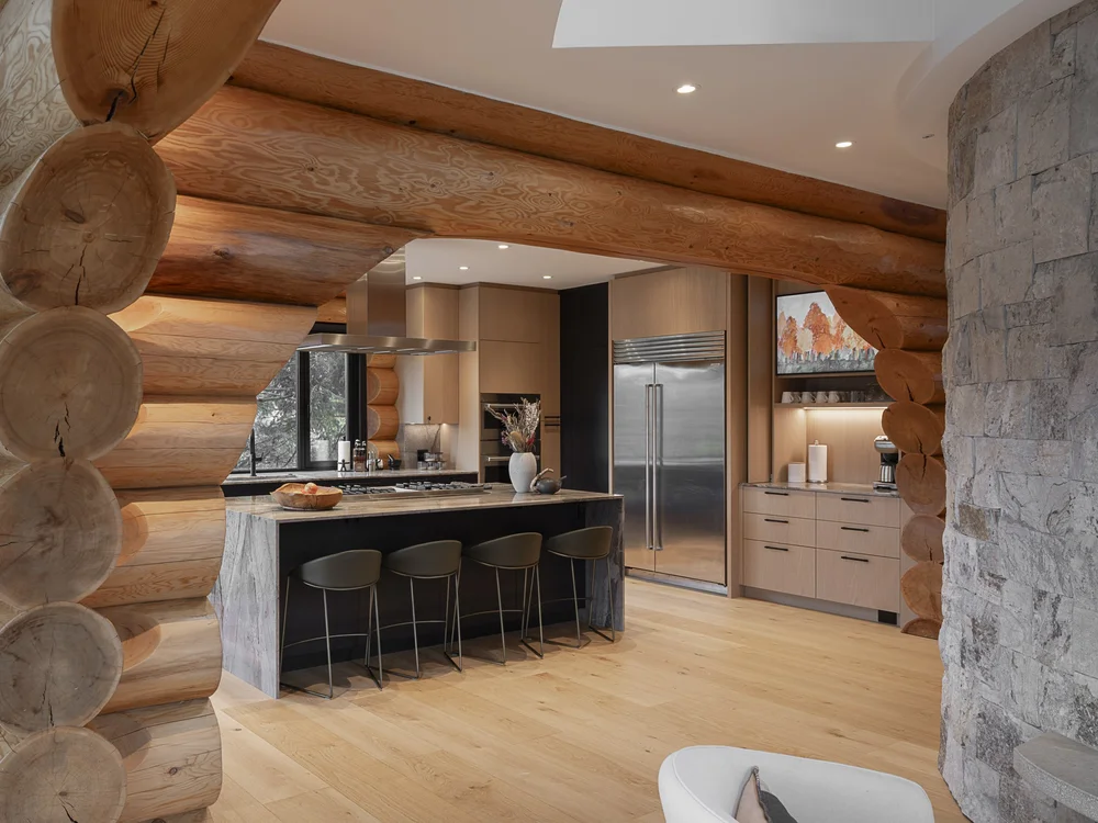 Looking past a stacked log corner into the kitchen, island and stools under a metal hood