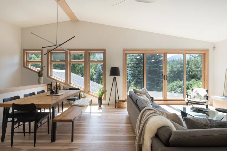 Dining table and sofa under a vaulted ceiling, wood framed windows onto forest and mountain