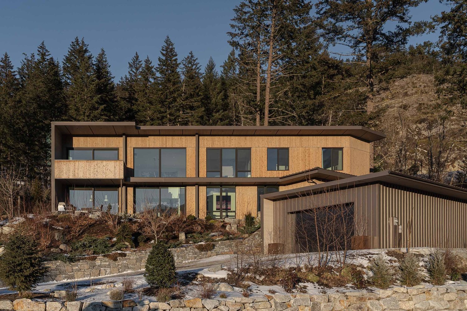 Two storey elevation in wood cladding above stone retaining walls, a low dark garage wing at right