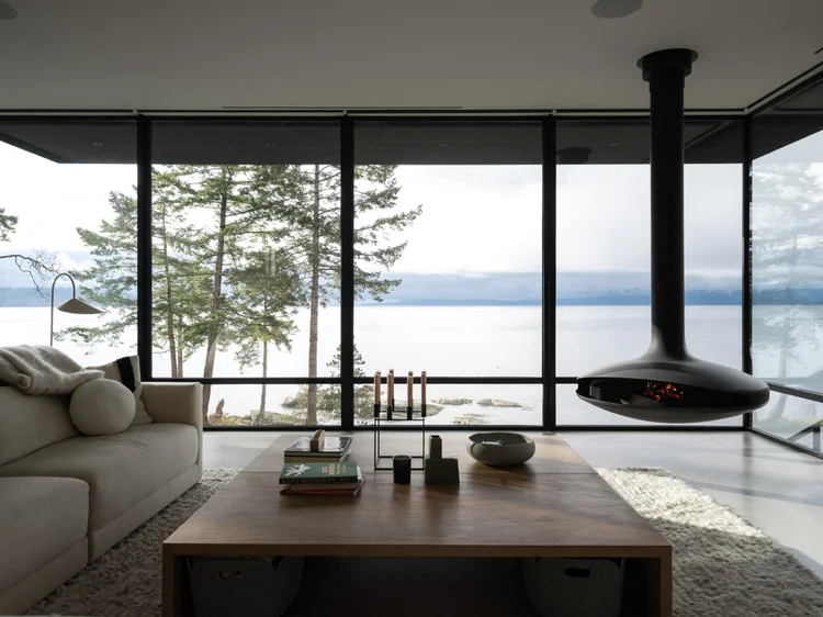 Low sofa and a wide table facing the water, the suspended fireplace burning at right