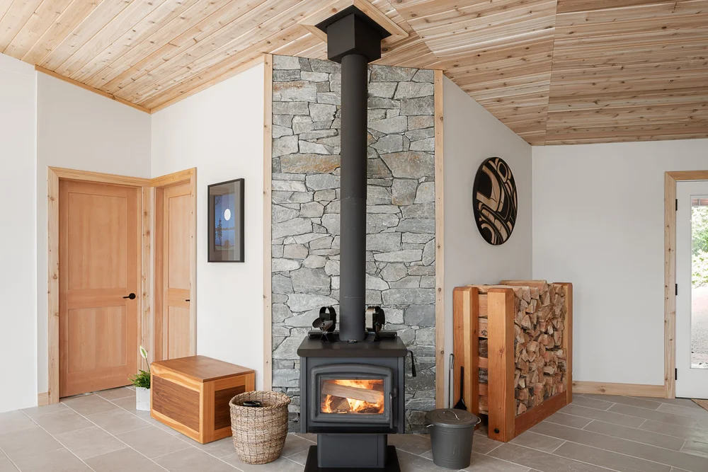 The wood stove alight on its stone hearth, split firewood stacked in a wood rack