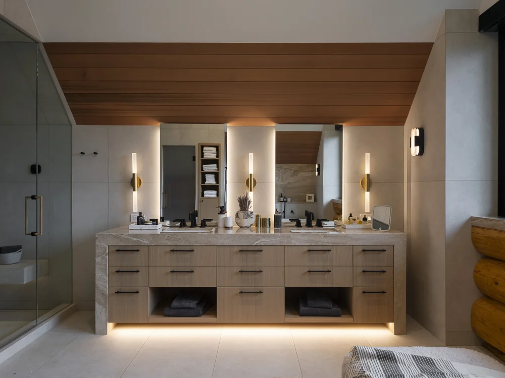 The vanity run under linear sconces, a shower at left and the room reflected in the mirror