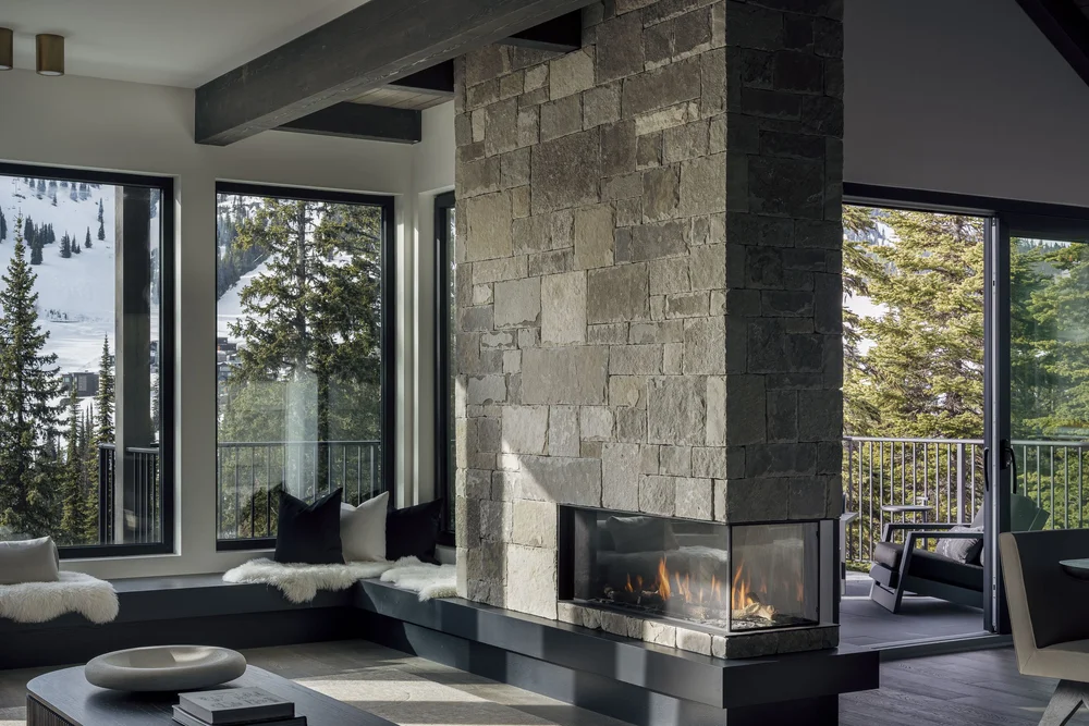 Stone chimney with a lit three sided fireplace, windows to snow on both sides