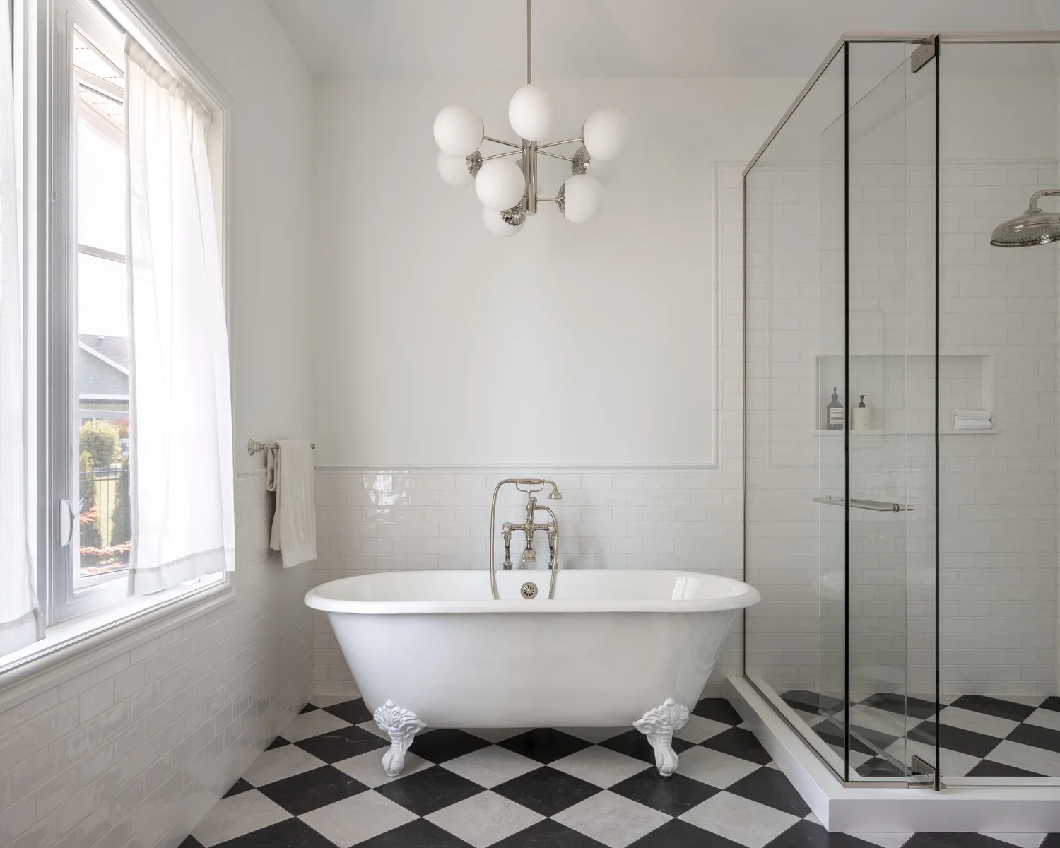 A clawfoot tub at a curtained window beside a glass shower, checkerboard floor underfoot