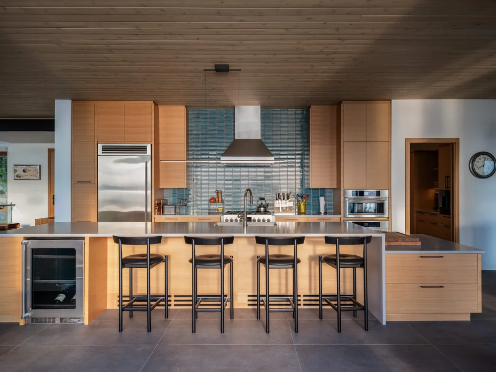 Kitchen island with four stools under a wood ceiling, ovens set into the cabinetry