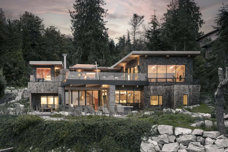 The house lit at dusk above a rock shoreline, low shingled volumes under wide overhangs