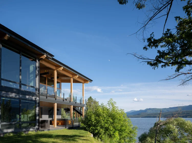 Glazed two storey elevation above a lawn, covered decks on timber posts, lake and mountains beyond