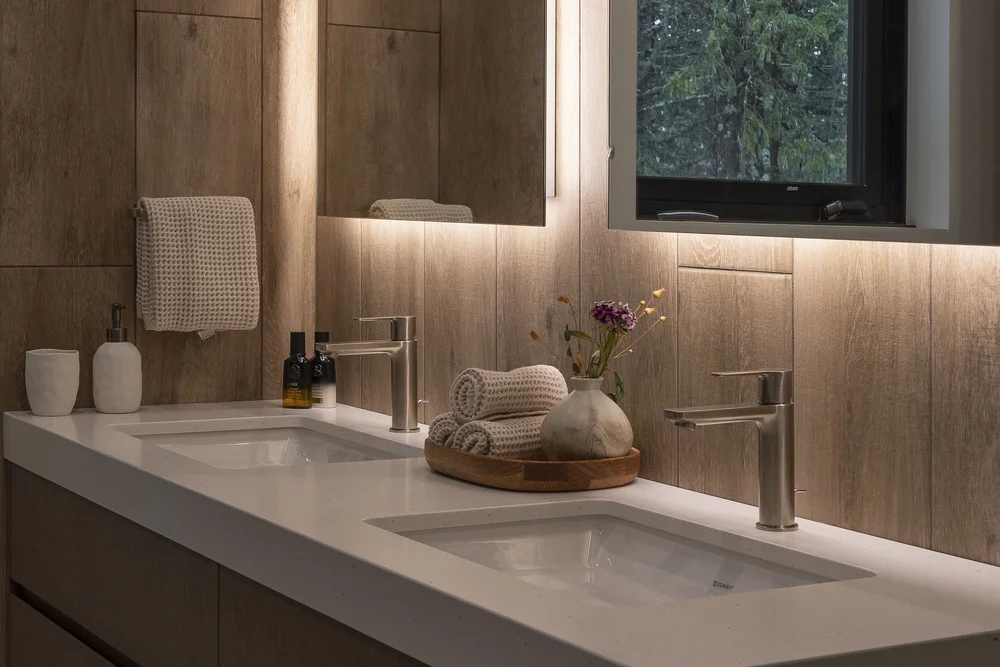 Twin basins in a pale counter under a backlit mirror, window onto dark conifers