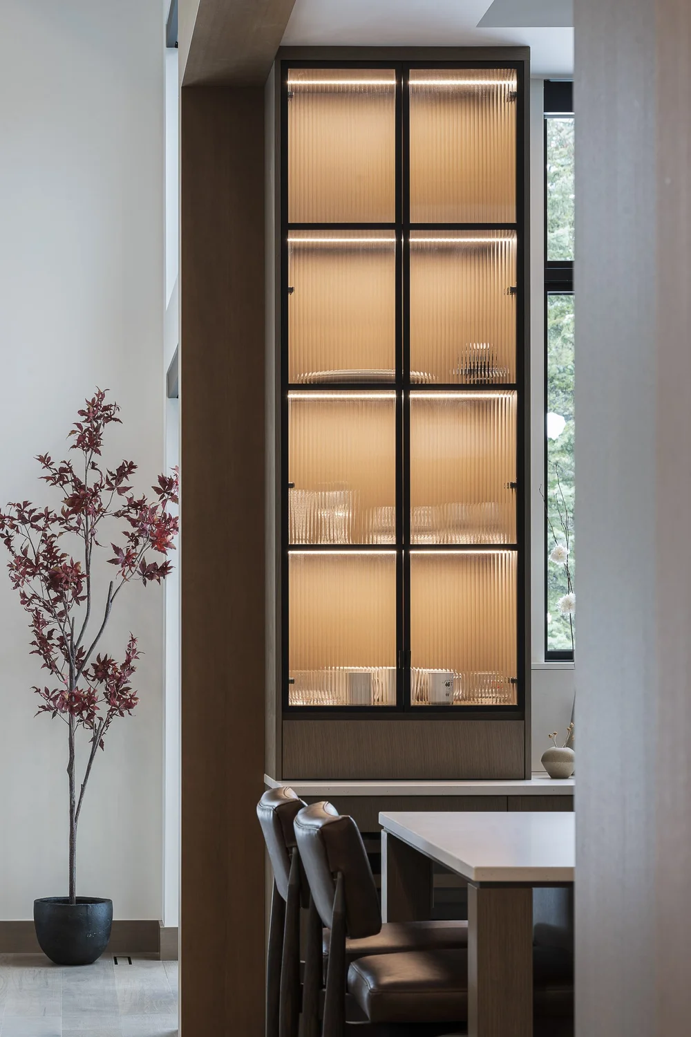 A tall lit display cabinet in a timber surround, dining chairs beyond