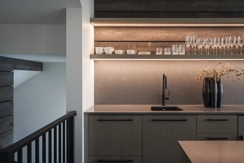 Bar counter with a lit open shelf of glassware above dark cabinets and a black tap