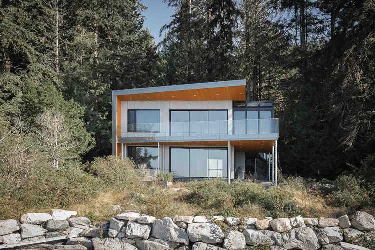 The house from below a rock wall, two storeys of glazing under a wood soffit, forest behind