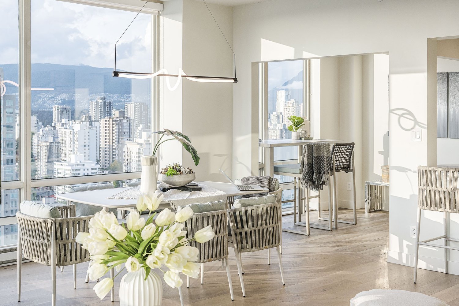Dining table set with white tulips under a linear pendant, the city through full height glass