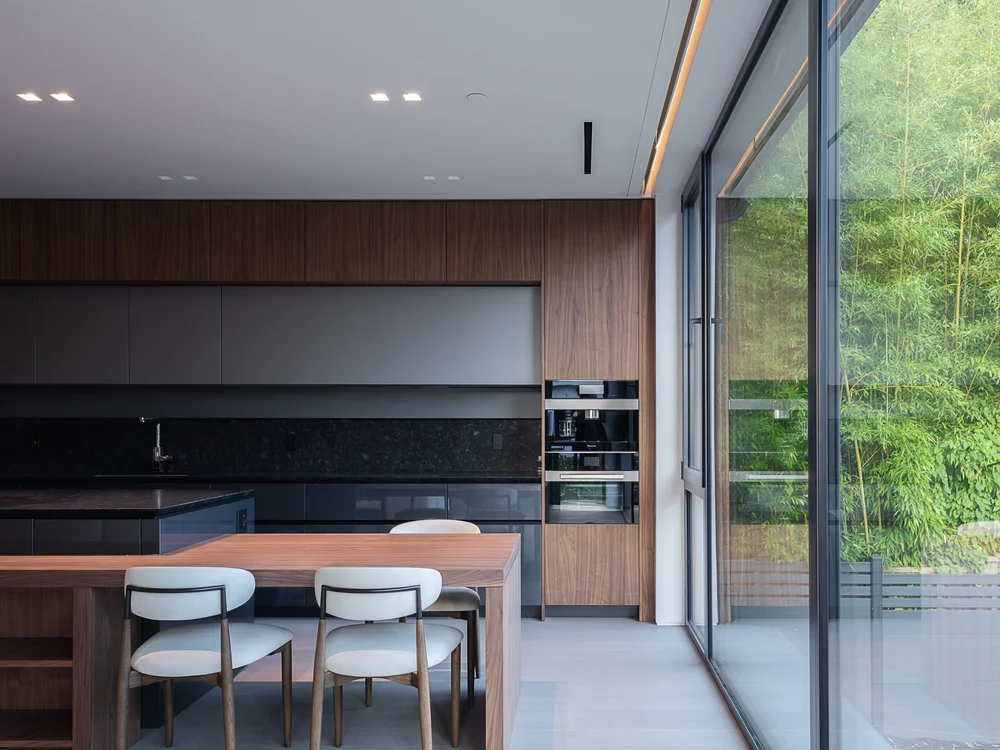 Kitchen in dark cabinetry with a stone counter, timber table and three chairs at the island