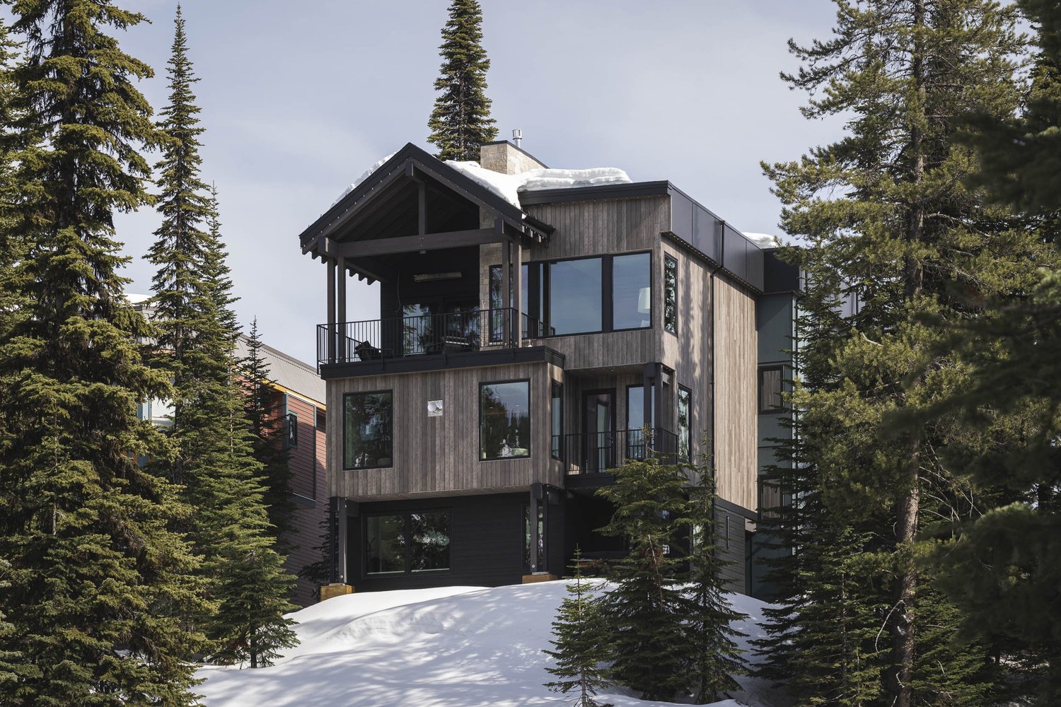 The house standing among tall evergreens above a deep bank of snow, dark and grey cladding