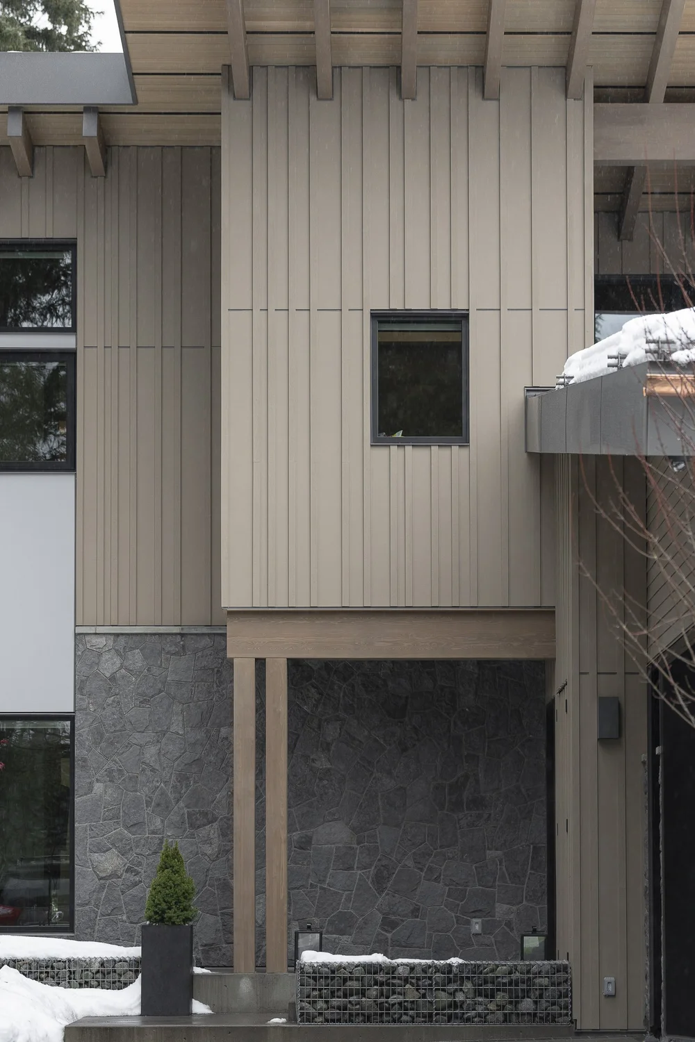Vertical siding above a dark stone base, a small square window, snow at the steps
