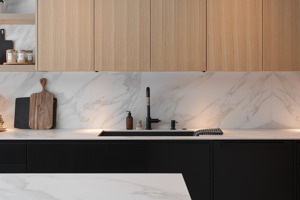 Sink run in dark cabinetry below a veined stone backsplash, a board propped behind