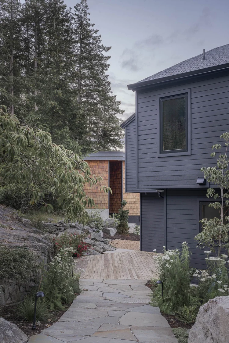 A dark clad house above a stone path, a shingled wing beyond and conifers behind