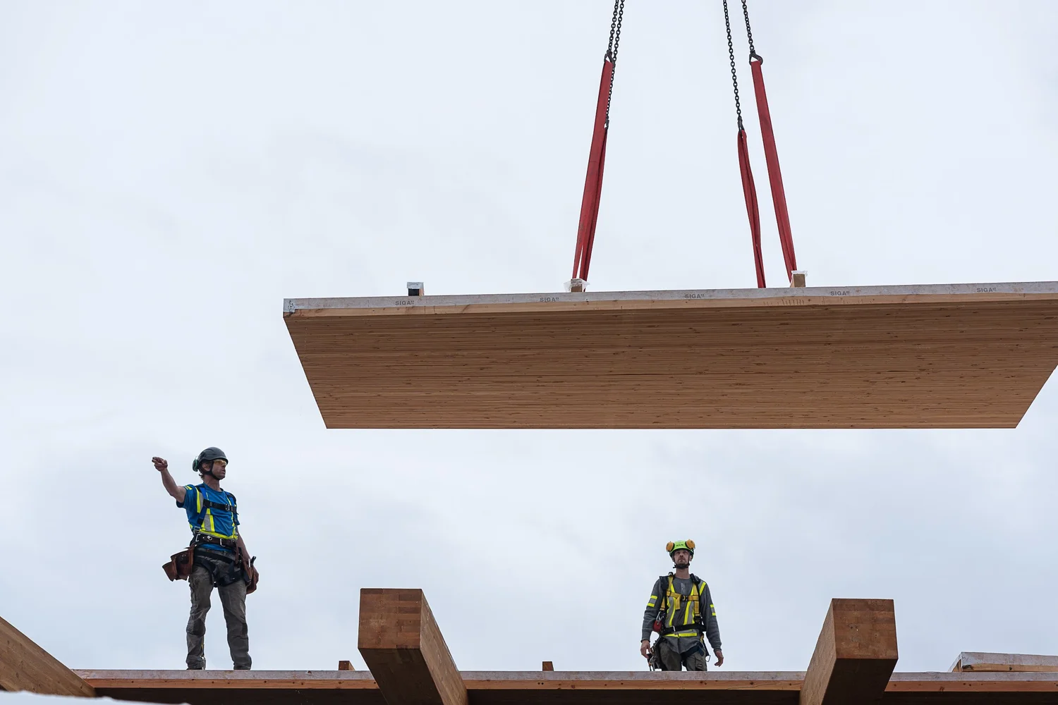 Workers on a mass timber beam structure with a CLT panel suspended by crane