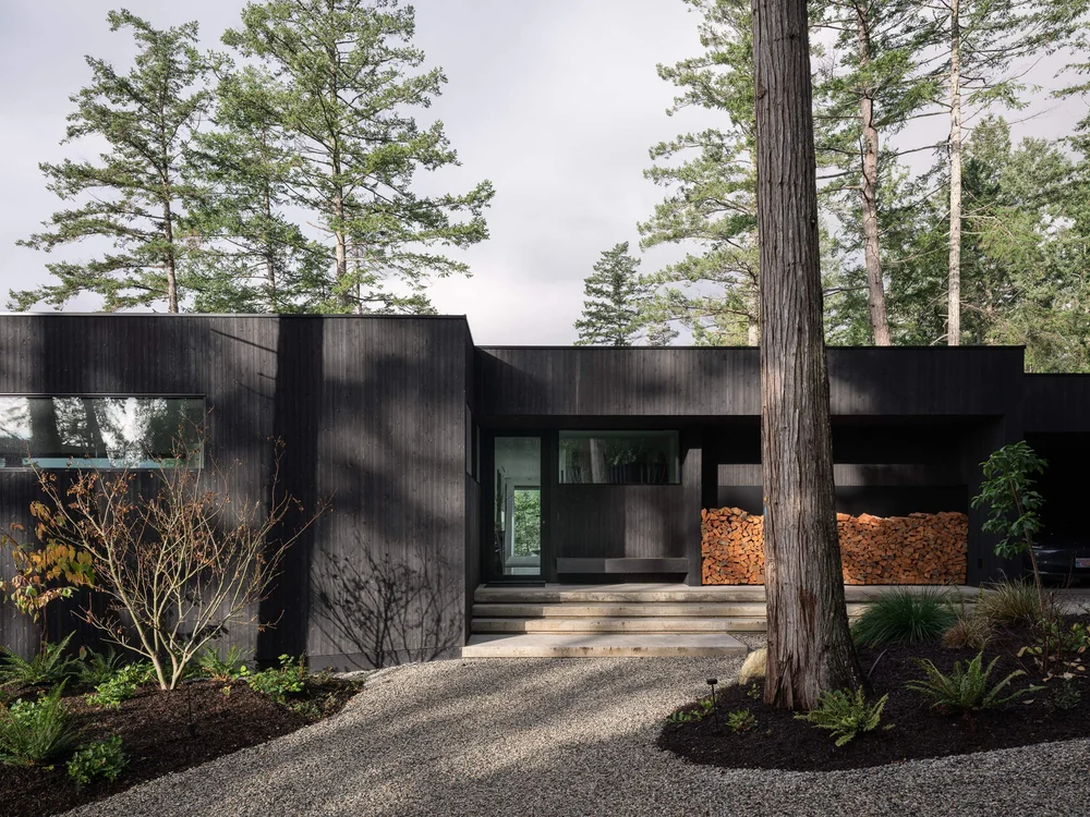 Gravel drive to a recessed entry, dark vertical cladding with firewood stacked beneath