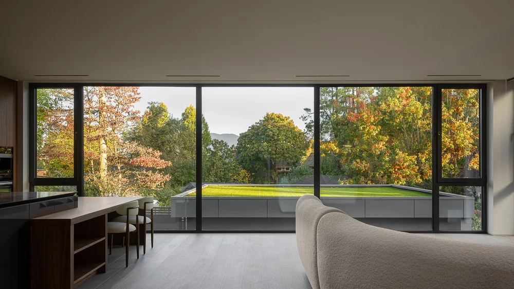 Full width glazing over the grass roof to autumn trees, kitchen table and sofa inside