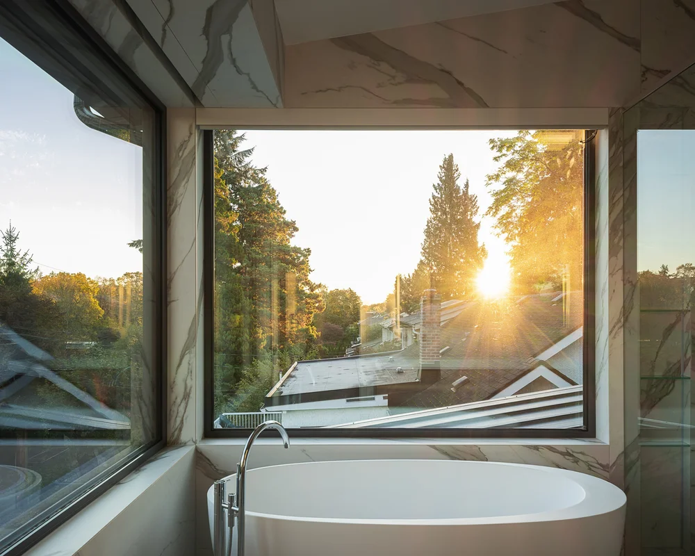 Freestanding tub at a corner window in veined stone, low sun through the trees