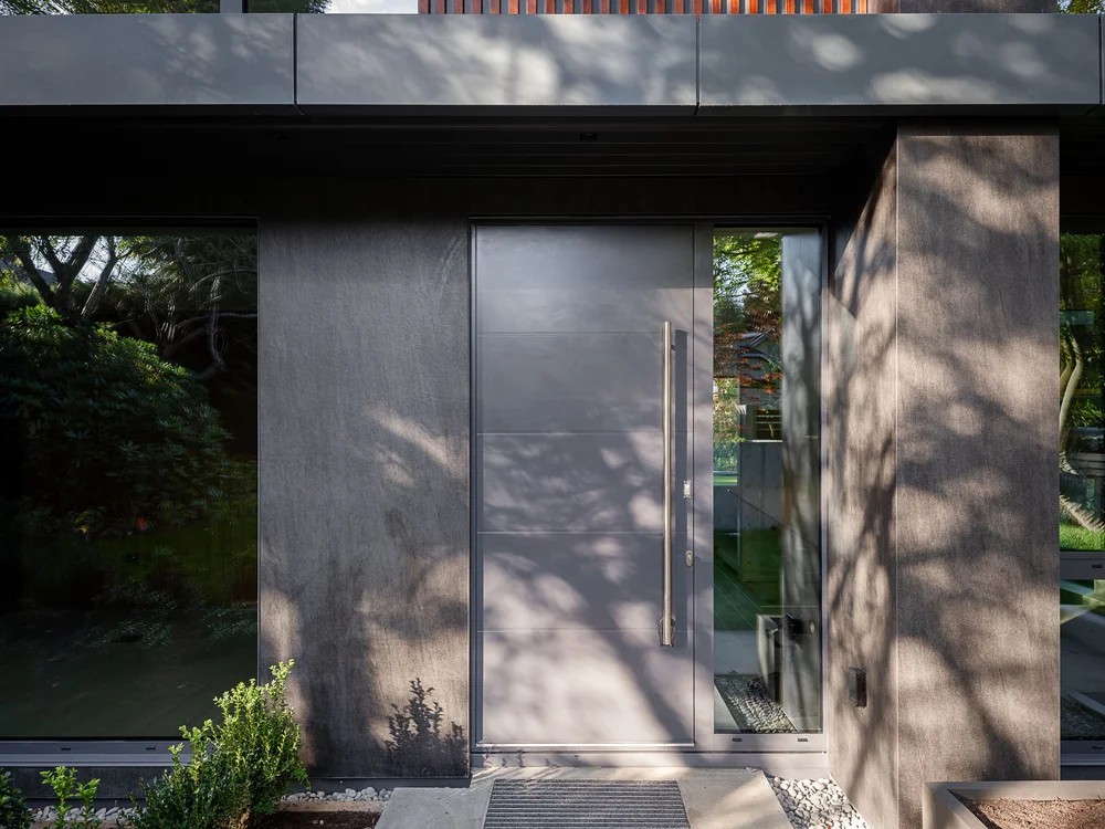 Entry door in dark panels under the cantilever, tree shadows across the wall