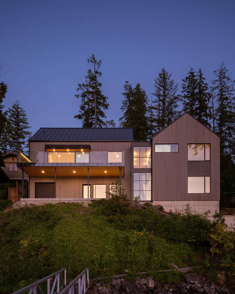 The house at twilight through the firs, windows lit along the elevation