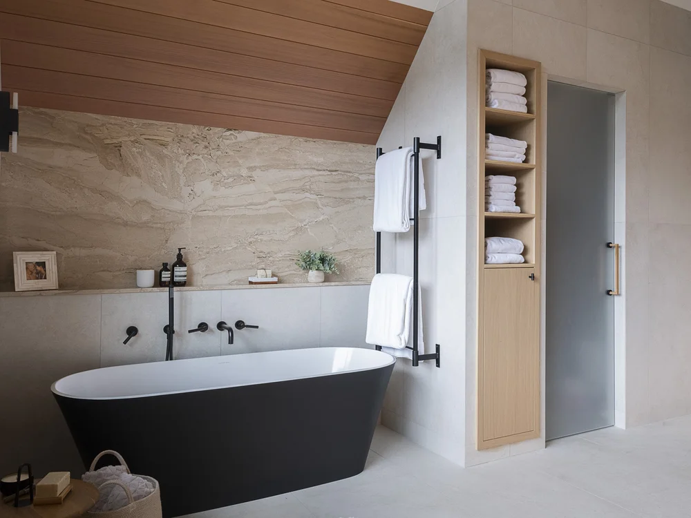 Freestanding black tub against a stone slab wall, towels stacked in a tall niche