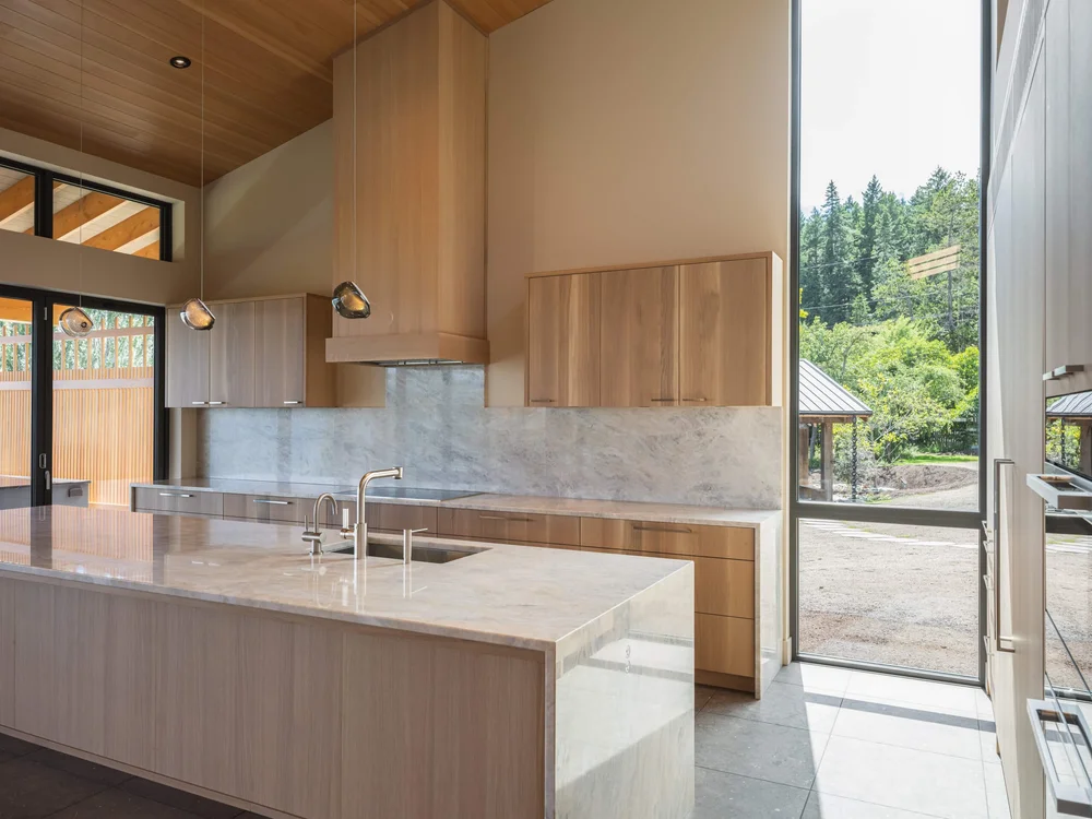Stone island under two pendants, the kitchen opening to glazing and the trees beyond
