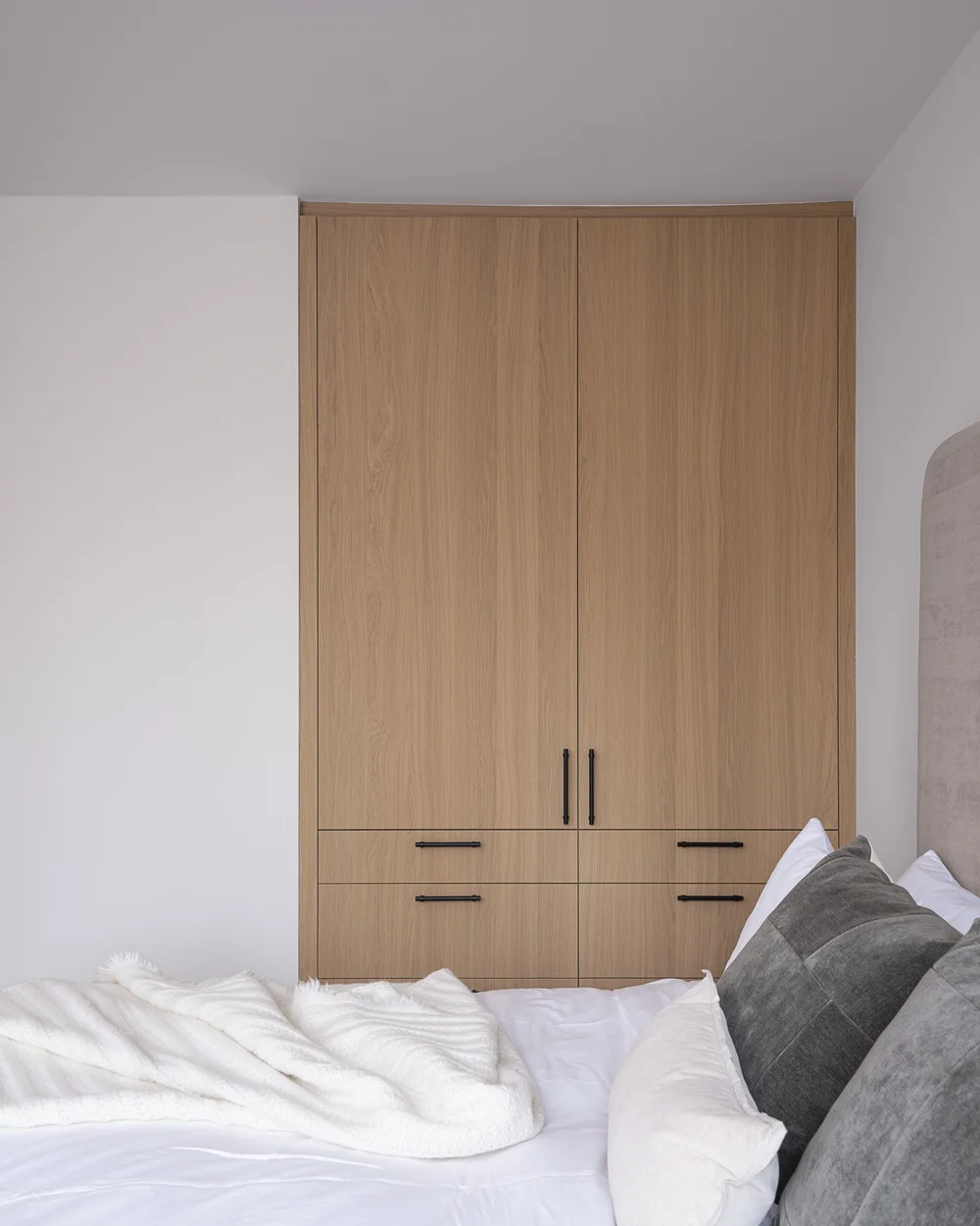 Bed made up in white and grey against a wall of pale wood wardrobes