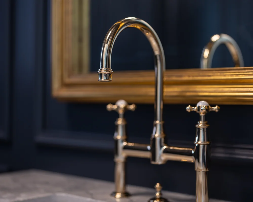 A gold toned bridge faucet against dark panelling, the corner of a gold frame behind