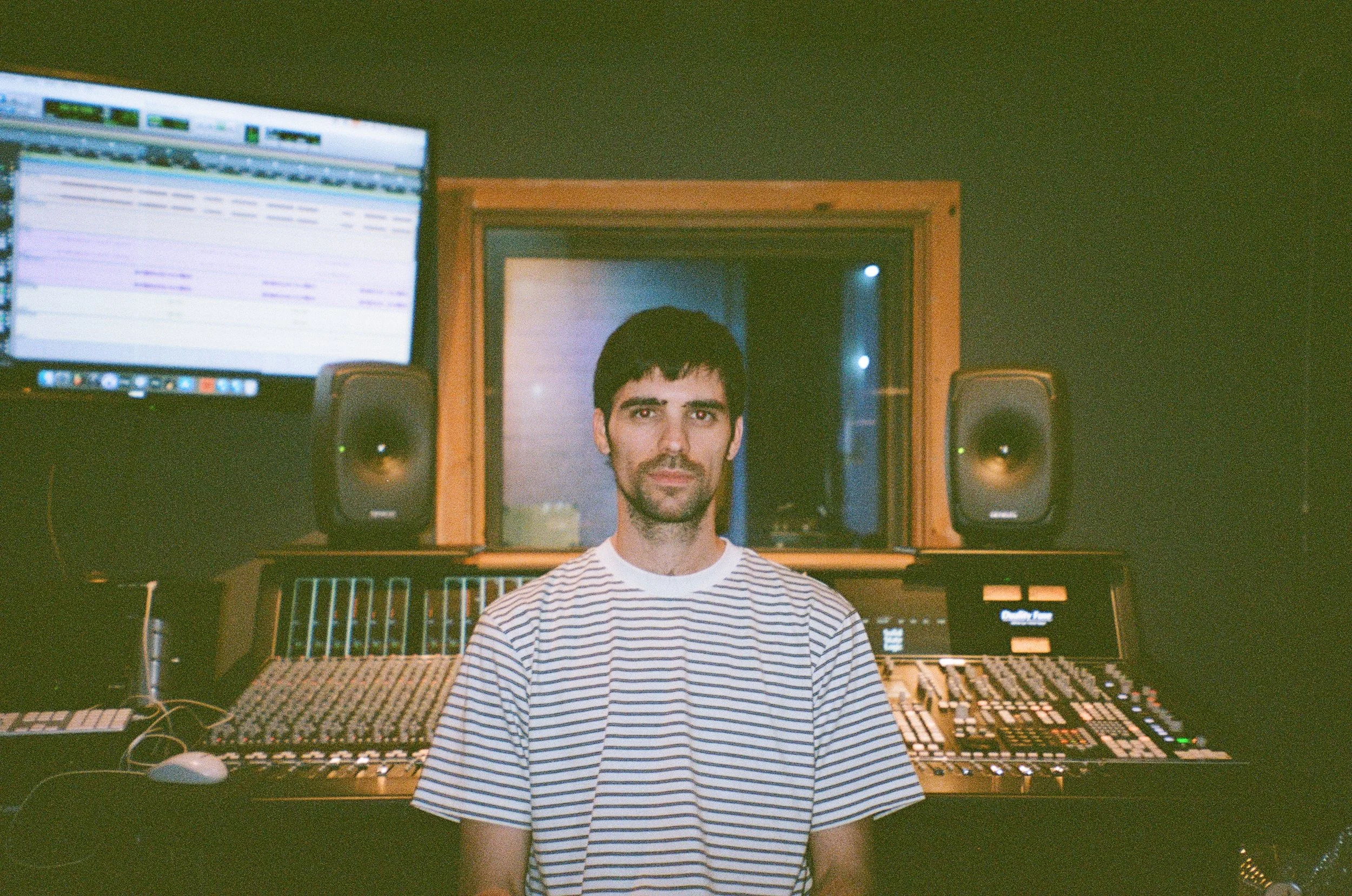 A man wearing a striped t-shirt sitting in front of a mixing console in a recording studio with music equipment, a computer screen, and audio speakers.