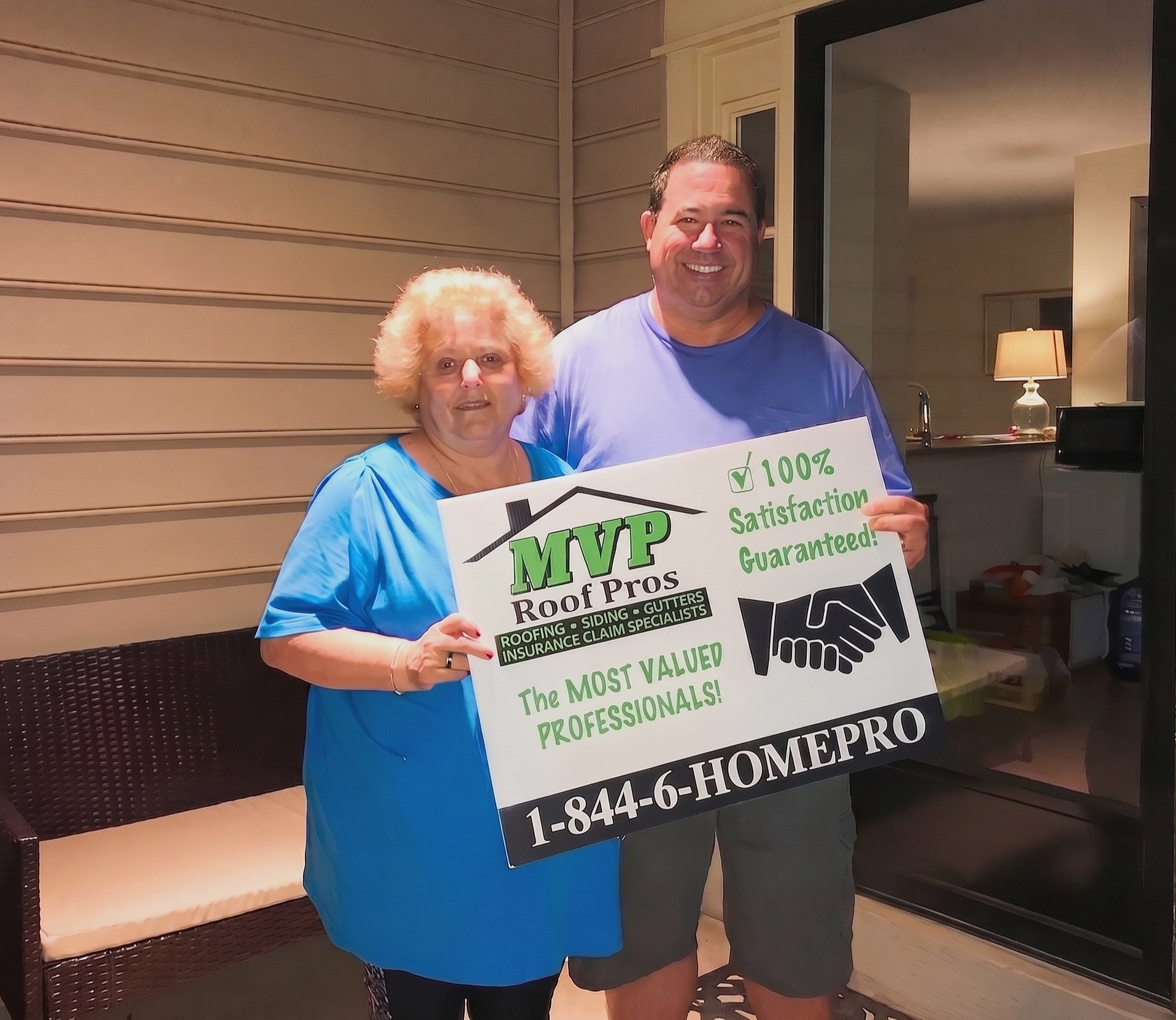 Two smiling people holding a sign promoting MVP Roof Pros services outside a house at night. The man is wearing a blue shirt and the woman is in a blue dress.